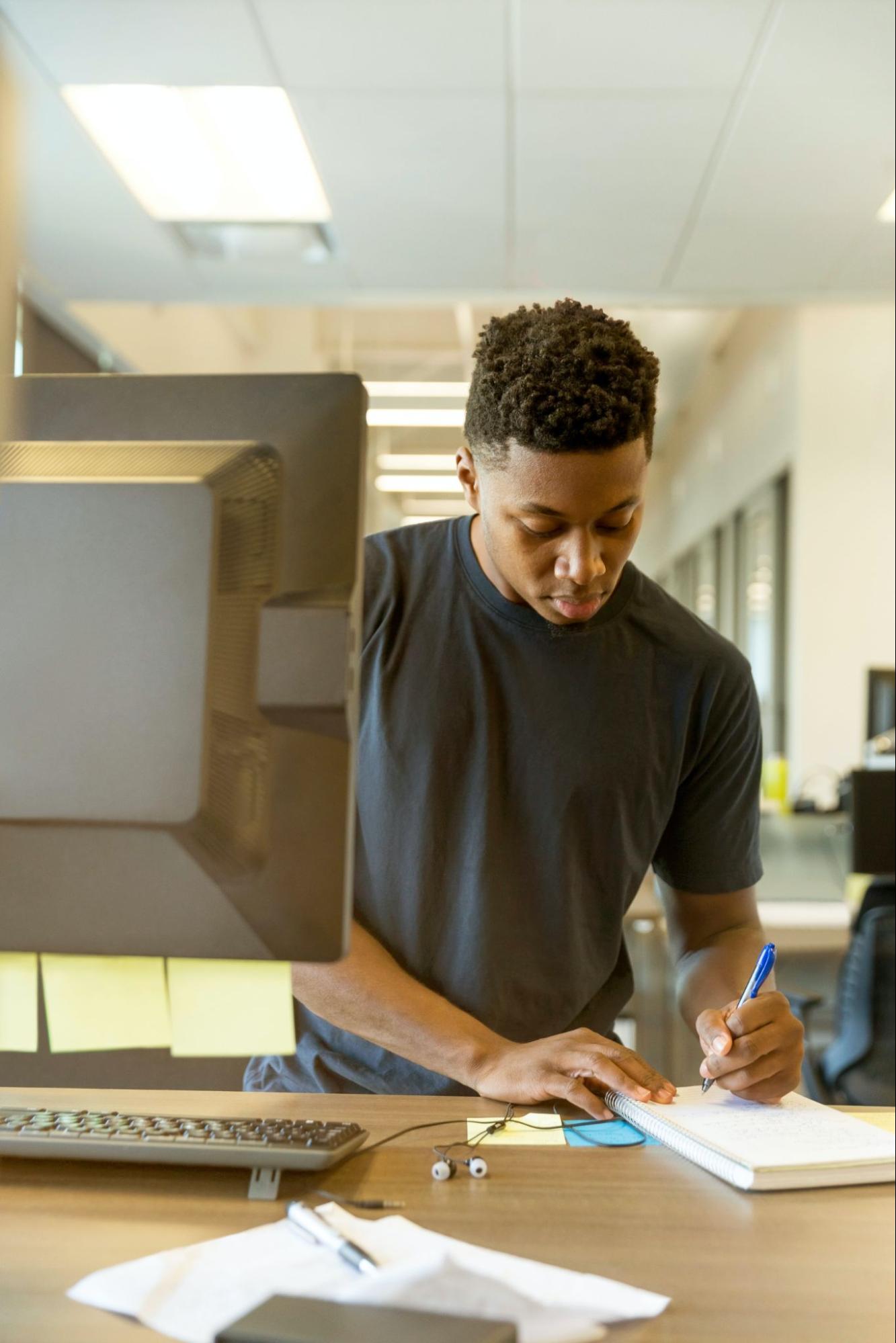 man writing on a notepad next to a computer