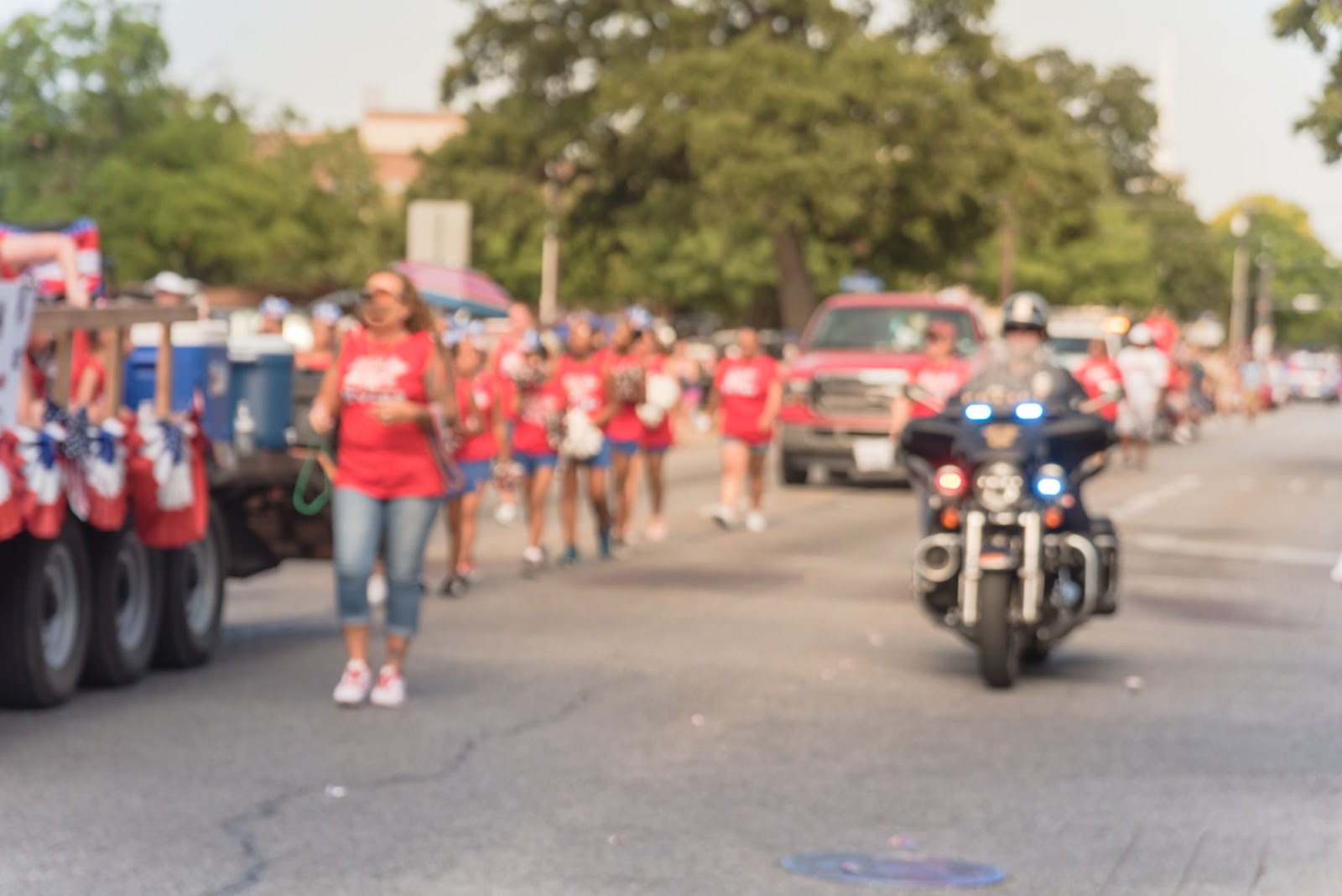 Blurry image of a fourth of July parade where everyone is wearing red shirts and blue pants while a policeman riding a motorcycle rides along side.