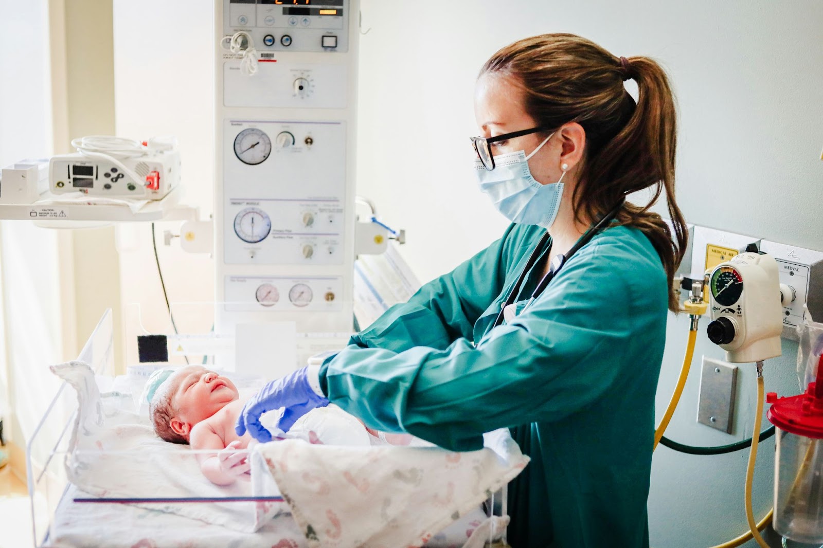 A female doctor or nurse wearing a gown, gloves, and mask to check an infants vitals. 