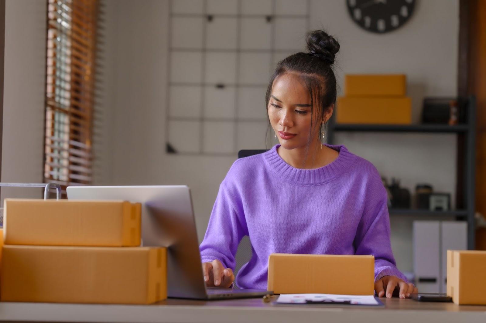Woman working on her computer at a desk in an office that seems to be set up for shipping prep.