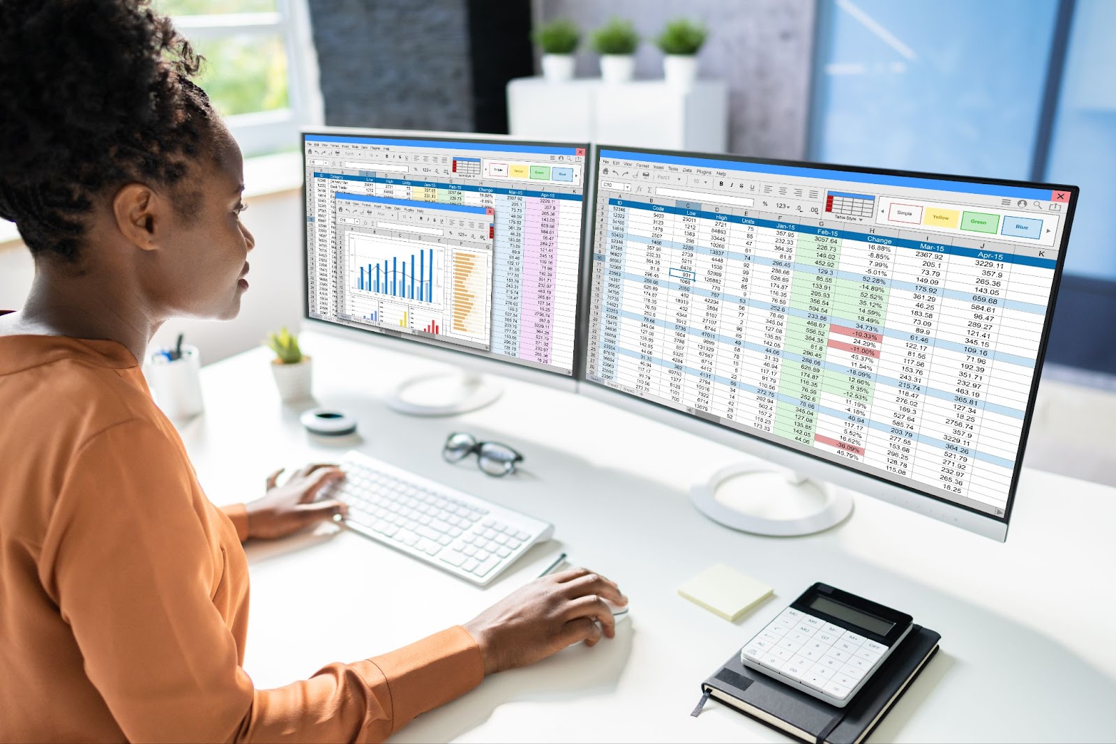 Woman in a very clean, naturally lit office with two large monitors - one showing data represented in a spreadsheet, the other showing data represented on a bar graph.