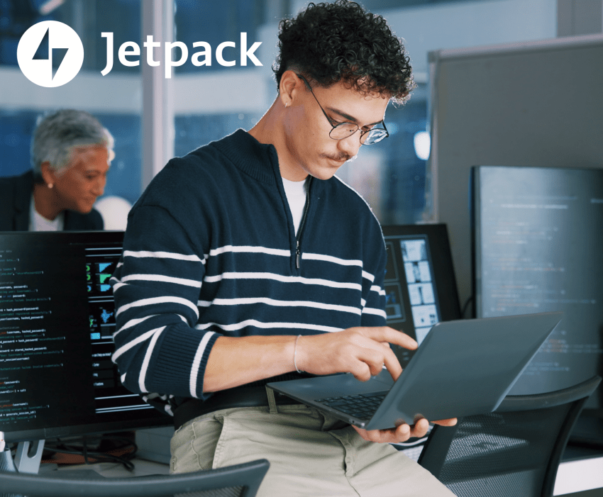 Man with glasses working on a laptop in an open office setting.